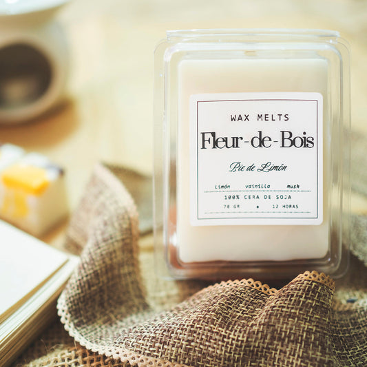 Un primer plano de "Wax Melts Barra de chocolate Fleur-de-Bois PIE DE LIMÓN" de Fleur-de-Bois, fundidos de cera de soja hechos a mano en un recipiente de plástico transparente, colocados sobre arpillera con un cuaderno borroso y una vela al fondo.