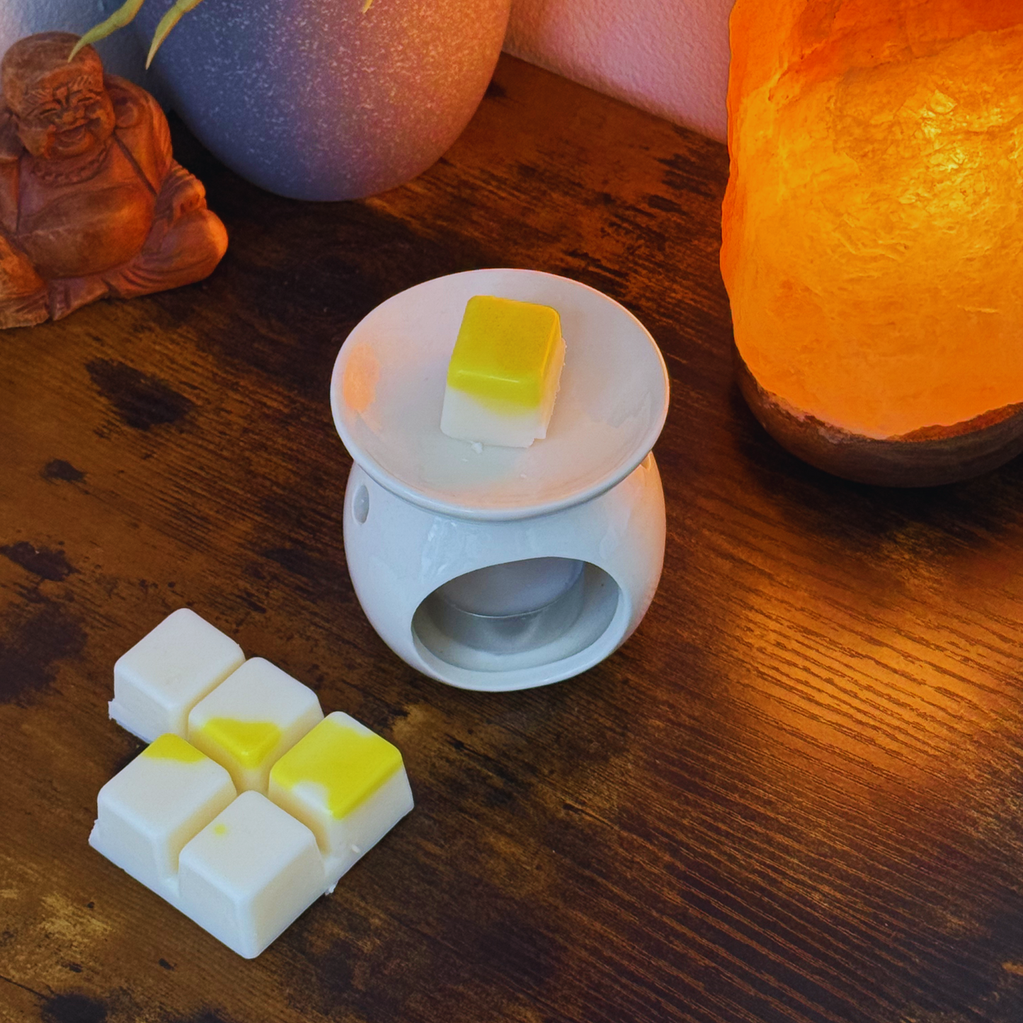 Un calentador de cera de cerámica blanca sobre una mesa de madera sostiene un wax melts Barra de chocolate de Cera de soja Fleur-de-Bois de chocolate PIE DE LIMÓN, con seis fundidos artesanales más cerca, además de una lámpara de sal naranja y una figurita de Buda al fondo.