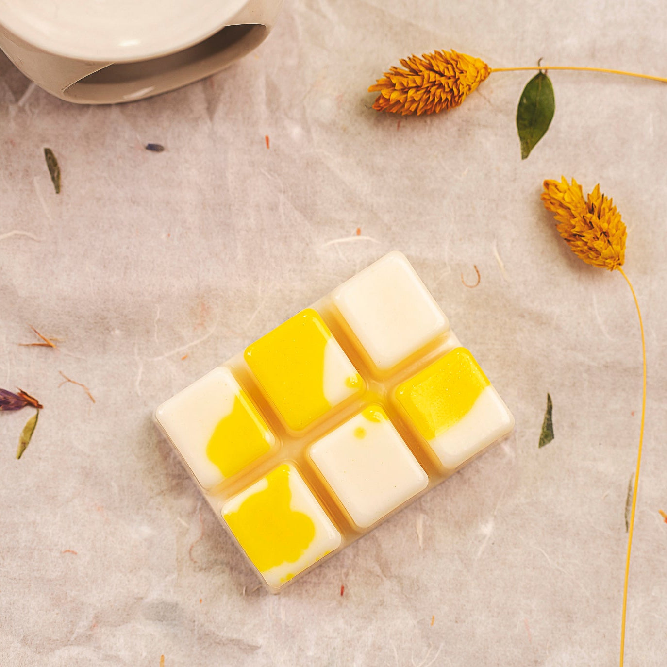 El Wax Melts Barra de chocolate Fleur-de-Bois Wax Melts PIE DE LIMÓN, una barra de estilo mosaico en blanco y amarillo inspirada en el chocolate, descansa sobre pergamino entre flores y hojas amarillas secas, con un cuenco de cerámica parcialmente visible en la esquina.