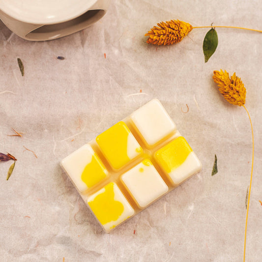 El Wax Melts Barra de chocolate Fleur-de-Bois Wax Melts PIE DE LIMÓN, una barra de estilo mosaico en blanco y amarillo inspirada en el chocolate, descansa sobre pergamino entre flores y hojas amarillas secas, con un cuenco de cerámica parcialmente visible en la esquina.