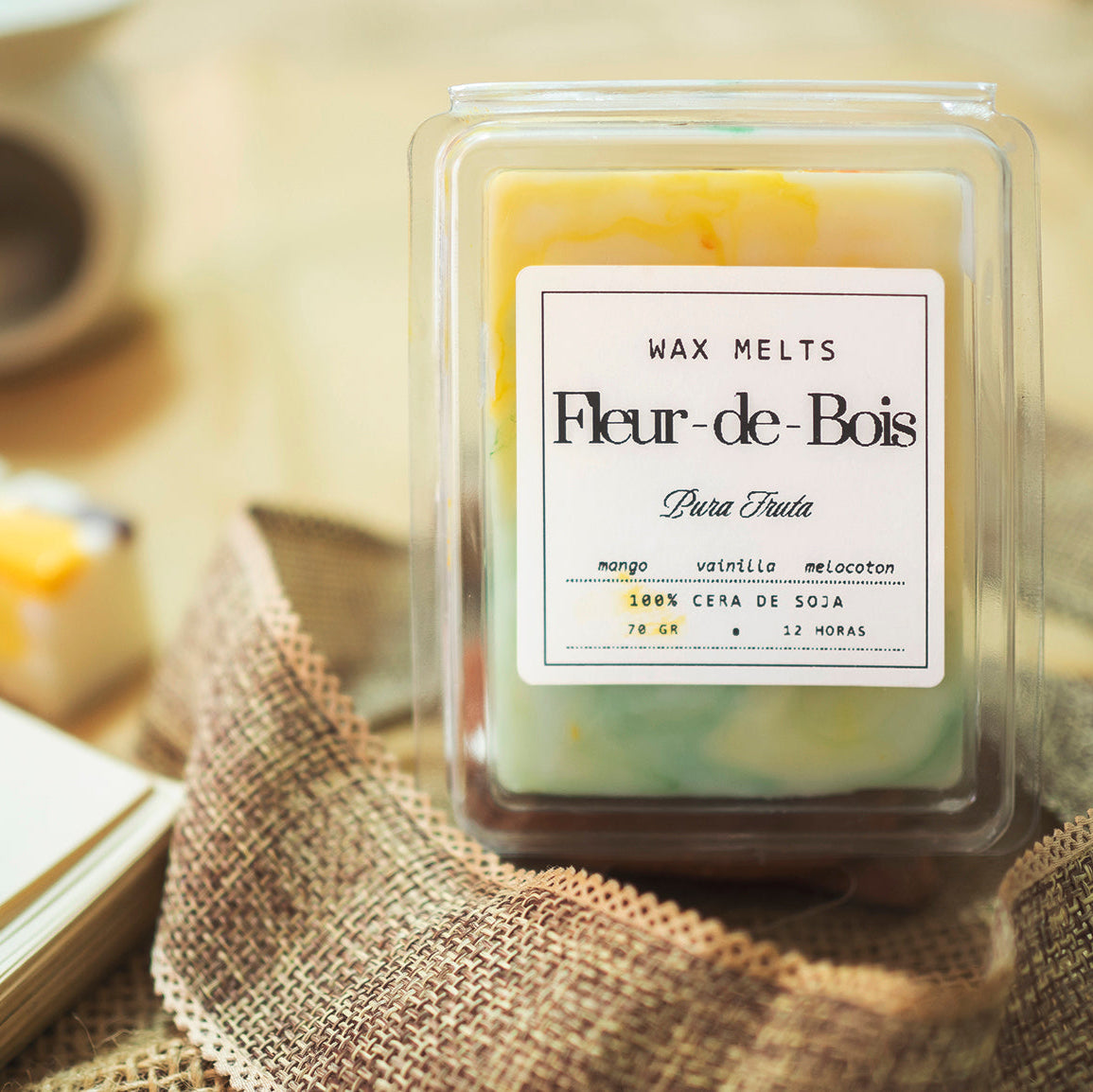 Un recipiente transparente con la etiqueta "Fleur-de-Bois, Wax Melts Barra de chocolate PURA FRUTA" sobre arpillera. Elaborados por Fleur-de-Bois a partir de cera de soja, estos fundidos combinan mango, vainilla y melocotón, un regalo personalizado ideal.