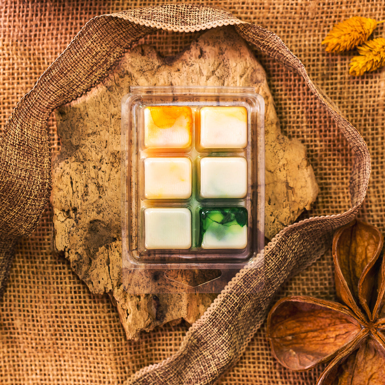 Un Wax Melts en forma de  Barra de chocolate Fleur-de-Bois PURA FRUTA de Fleur-de-Bois descansa sobre una corteza entre arpillera, cinta, hojas secas y flores amarillas, creando una escena rústica de otoño ideal como regalo personalizado.