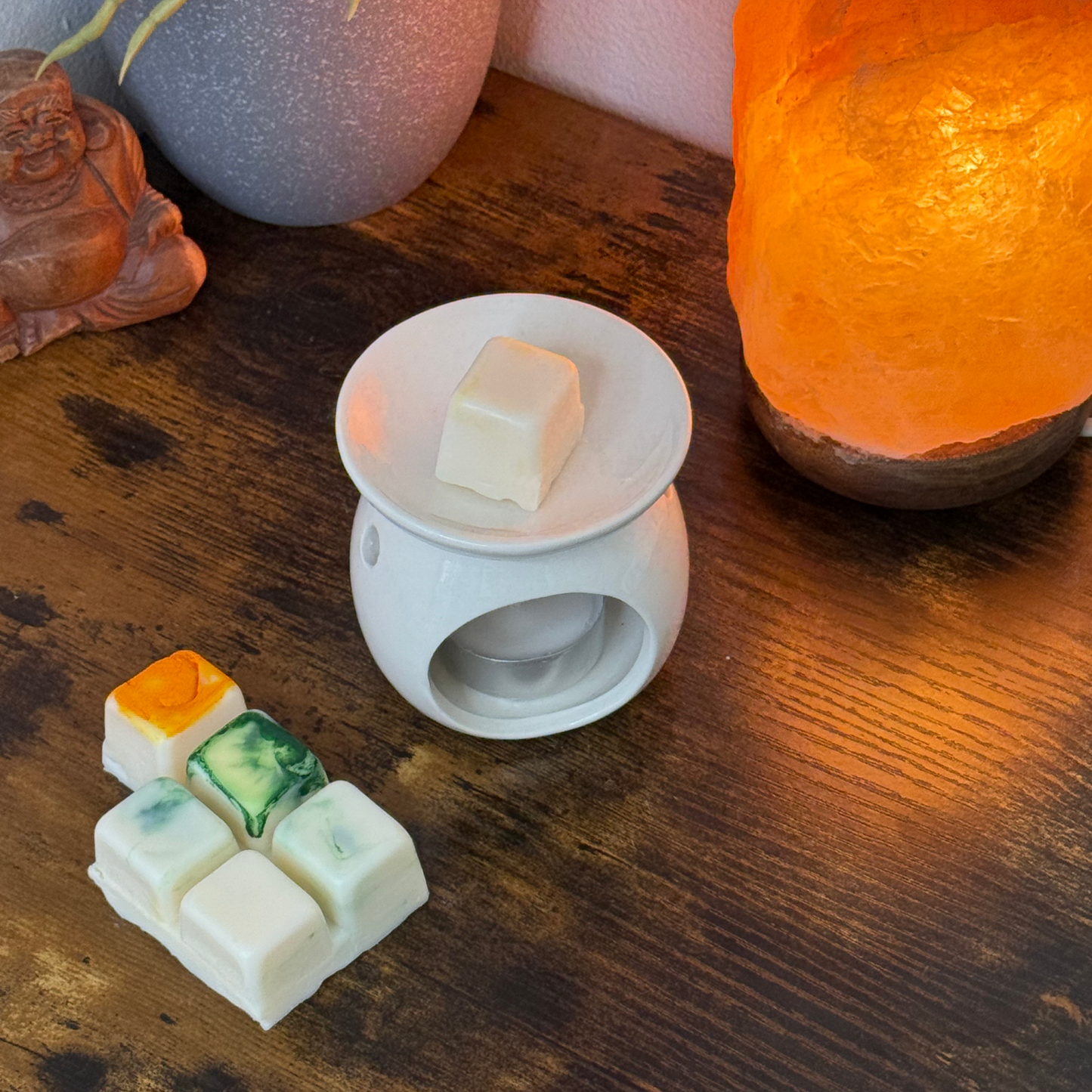 Sobre una mesa de madera hay un calentador de cera de cerámica blanca con un trozo de wax melts Barra de chocolate Fleur-de-Bois PURA FRUTA Wax Melts. Cerca hay coloridos fundidos Fleur-de-Bois hechos a mano y una lámpara de roca naranja brillante, con parte de una estatuilla de Buda en la esquina.