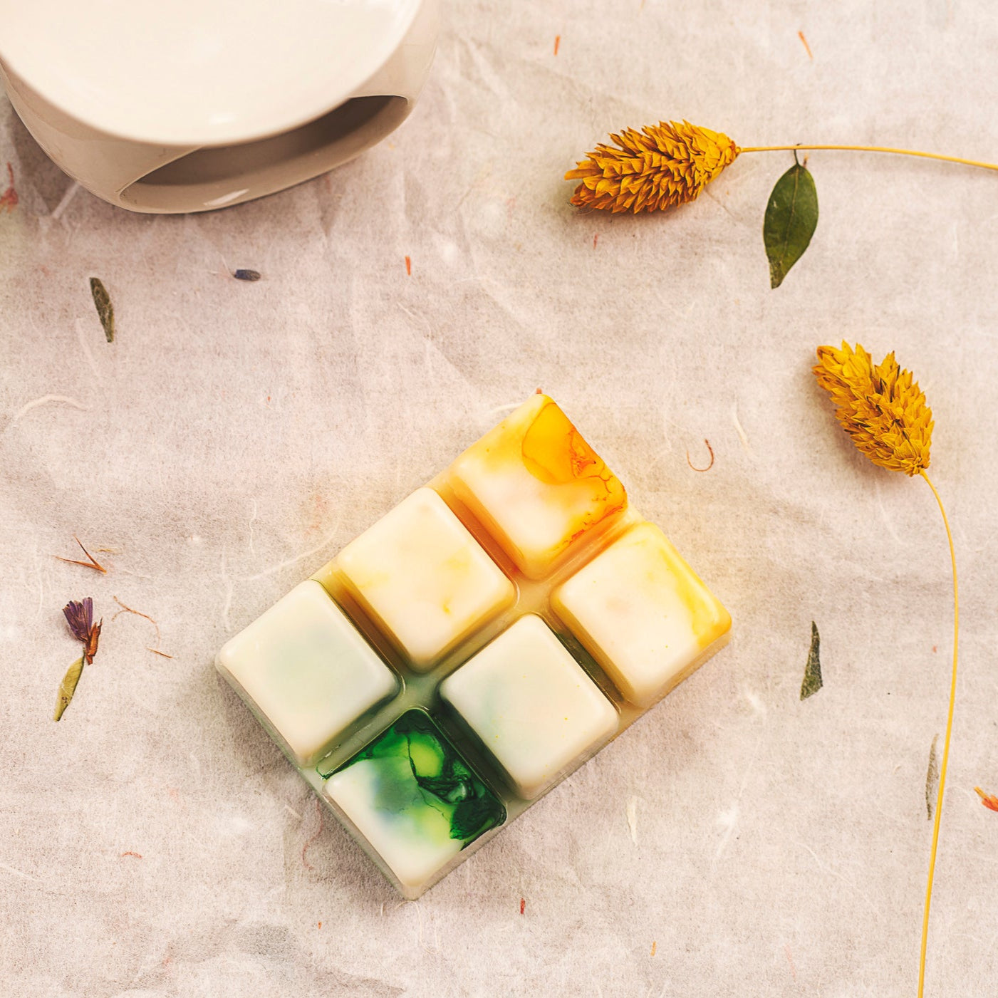 Un Wax Melts Fleur-de-Bois de forma Barra de chocolate PURA FRUTA, con seis gajos en una base blanca con remolinos verdes, amarillos y naranjas, descansa sobre una superficie de textura ligera junto a flores amarillas secas y hojas esparcidas.