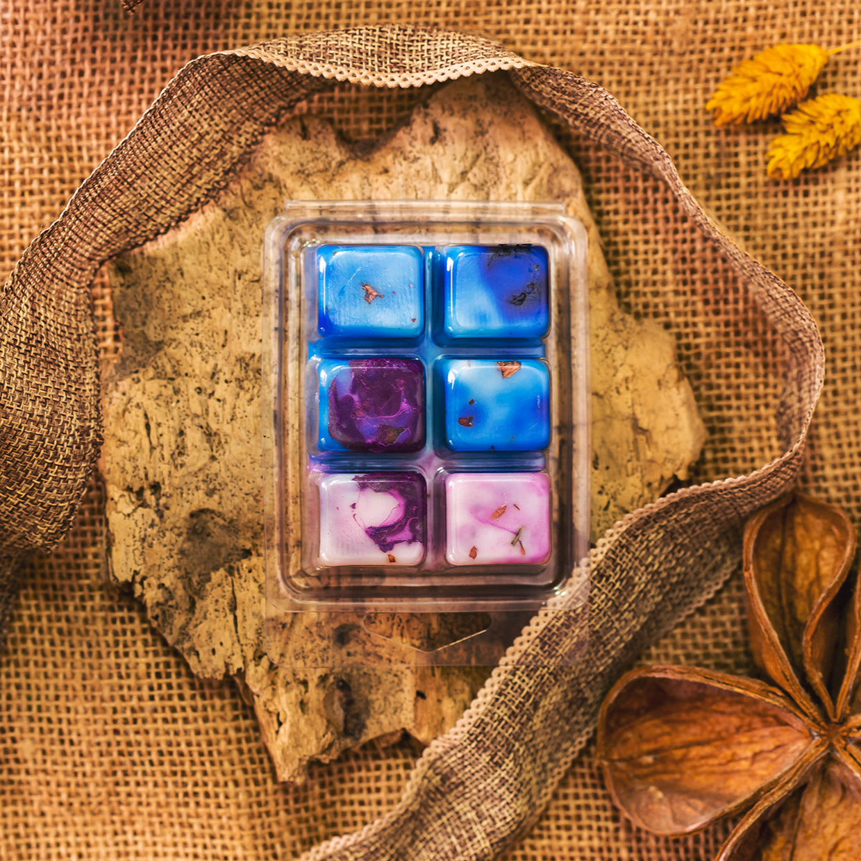 Un recipiente de plástico contiene seis coloridas Fleur-de-Bois Wax Melts Barra de chocolate PURPLE WOOD en azul, morado y blanco sobre un trozo de corteza con arpillera, cinta y flores secas: un regalo ideal y personalizado.