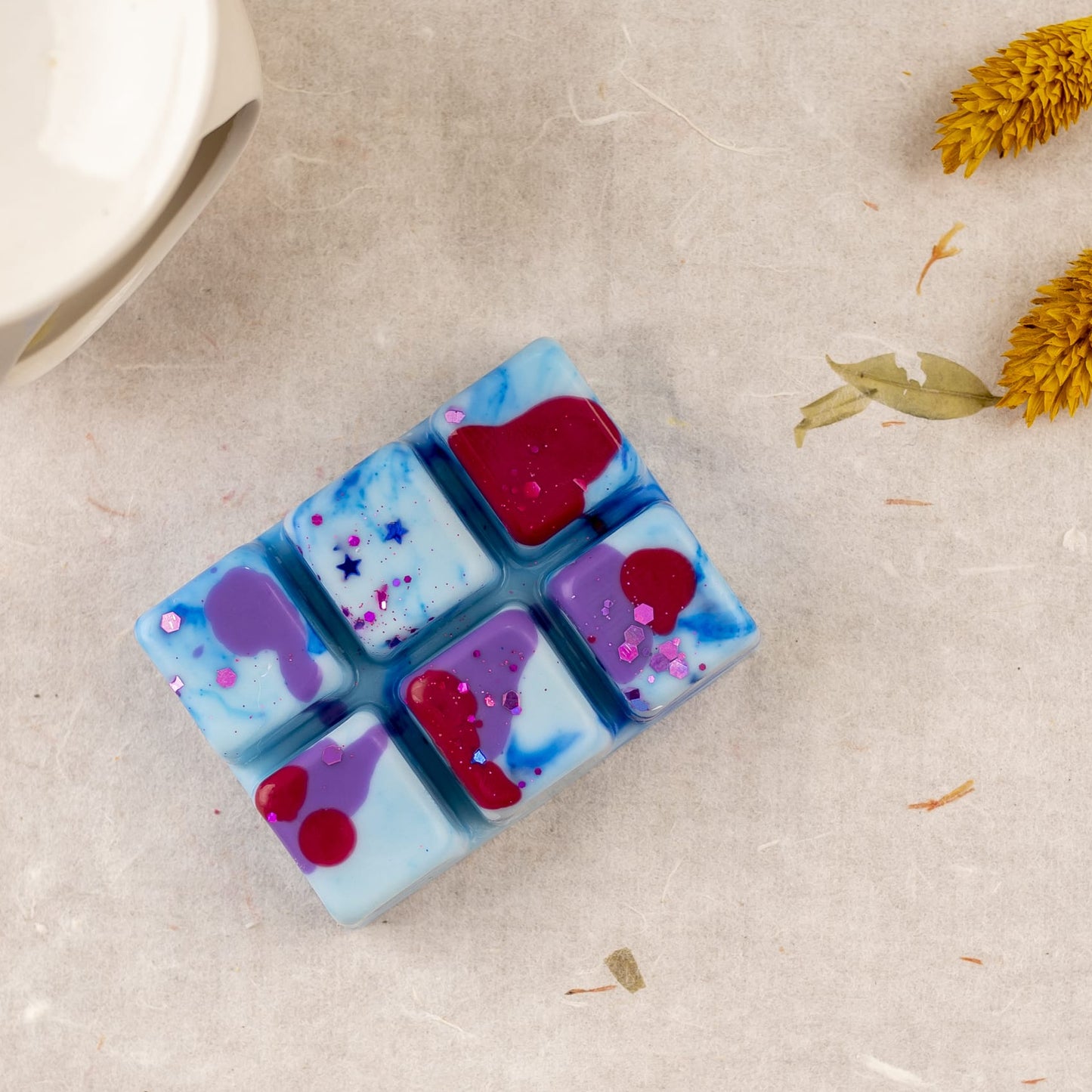 Wax Melts azul, morado y rojo de Fleur-de-Bois, la "Barra de chocolate Wax Melts Fleur-de-Bois PURPLE WOOD", adornados con flores secas, descansan sobre pergamino: un regalo personalizado ideal para cualquier ocasión.