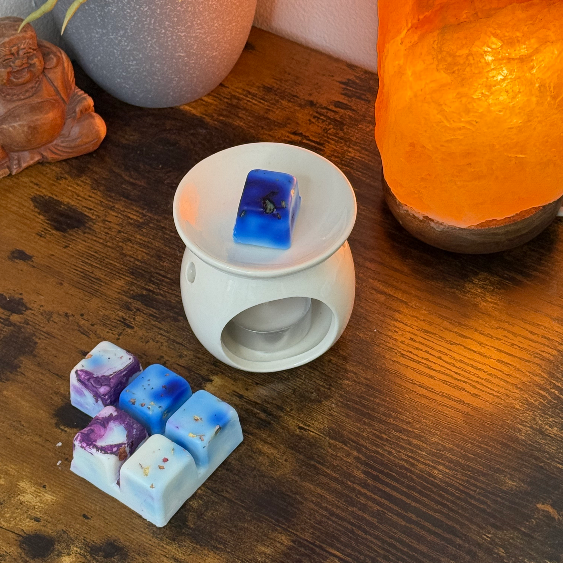 Un calentador de cera blanco con una barra de chocolate Fleur-de-Bois Wax Melts PURPLE WOOD sobre una superficie de madera. Cerca hay una variedad de cera de soja derretida, una lámpara de sal del Himalaya, una estatua de Buda y una maceta gris, ideal para un regalo personalizado.
