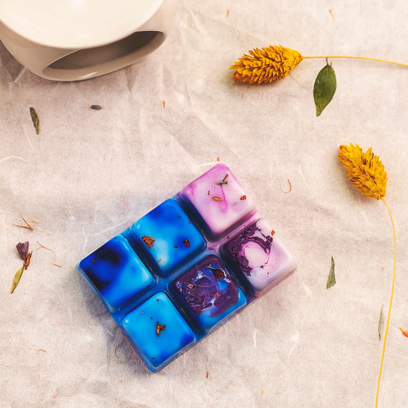 Seis vibrantes Wax Melts azules y morados de Fleur-de-Bois, la "Barra de chocolate Wax Melts Fleur-de-Bois PURPLE WOOD", adornados con flores secas, descansan sobre pergamino: un regalo personalizado ideal para cualquier ocasión.