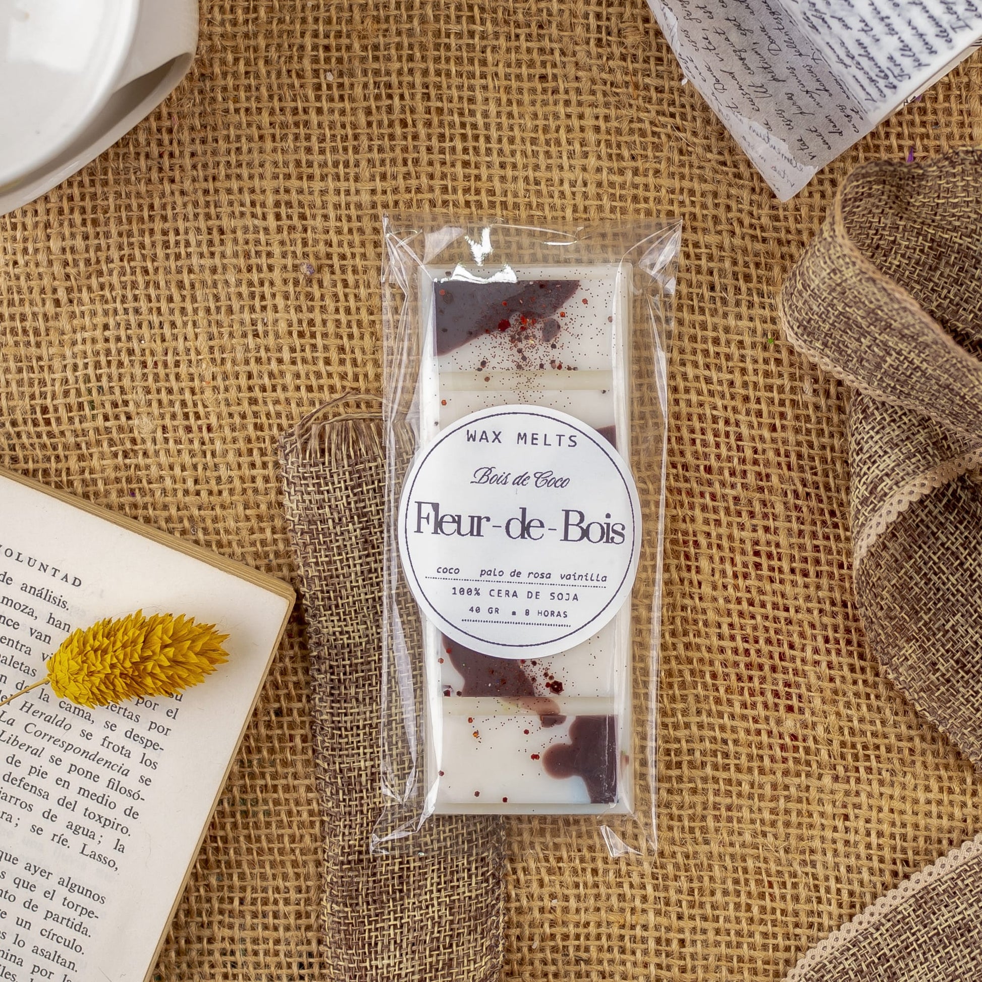 Un paquete de Melts de Cera Láminas Aromáticas Fleur-de-Bois BOIS DE COCO de Fleur-de-Bois está colocado sobre arpillera, rodeado de un libro, una flor amarilla seca, papel enrollado y una cinta marrón. Los fundentes blancos presentan motas marrones y rojas para unas fragancias elegantes.