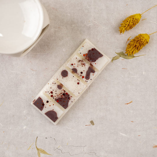 Fleur-de-Bois Wax Melts Láminas Aromáticas BOIS DE COCO, que se asemejan a un elegante chocolate blanco con salpicaduras de bayas y pequeñas coberturas, se exponen sobre una superficie texturizada junto a dos flores amarillas secas y un plato de cerámica blanca.