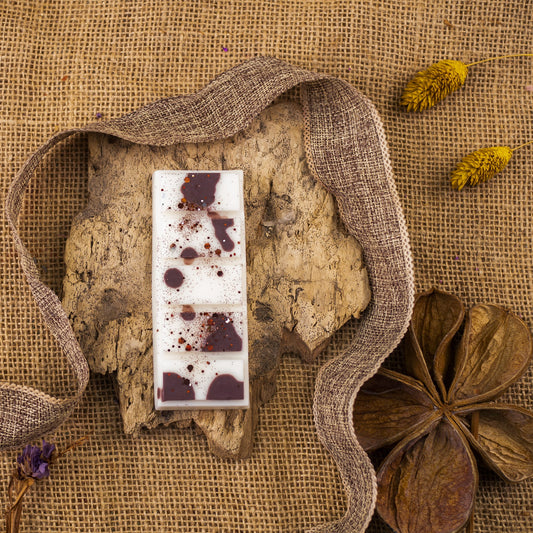 Las Wax Melts de Fleur-de-Bois Láminas Aromáticas BOIS DE COCO, diseñadas con detalles oscuros y espolvoreados, parecen una elegante confitería mientras descansan sobre corteza, rodeadas de flores secas y cinta sobre tela de arpillera rústica.