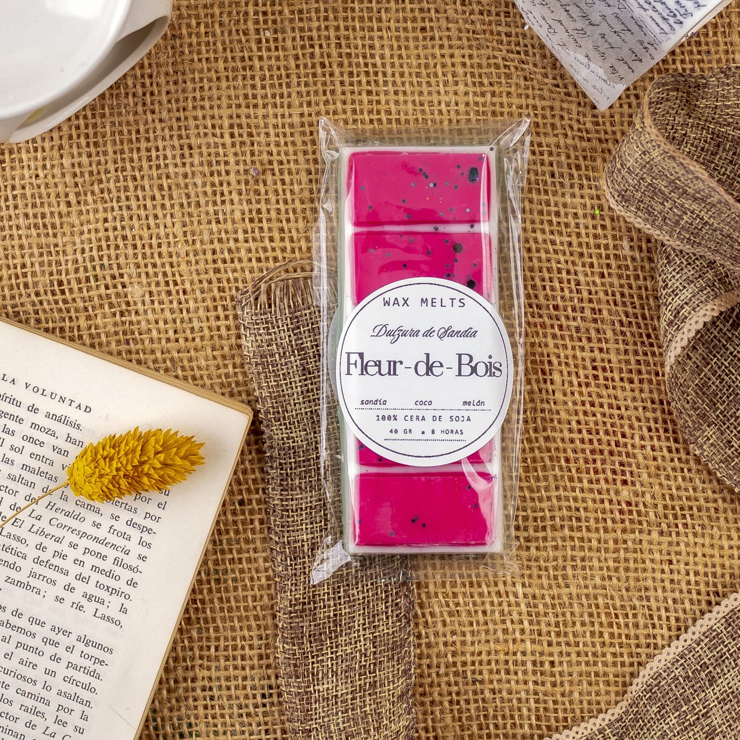 Un paquete de Láminas Aromáticas Fleur-de-Bois DULZURA DE SANDÍA de Fleur-de-Bois descansa sobre arpillera, rodeado de un libro abierto, papel, un cuenco de cerámica y una flor seca amarilla, perfecto para disfrutar de fragancias elegantes en casa.