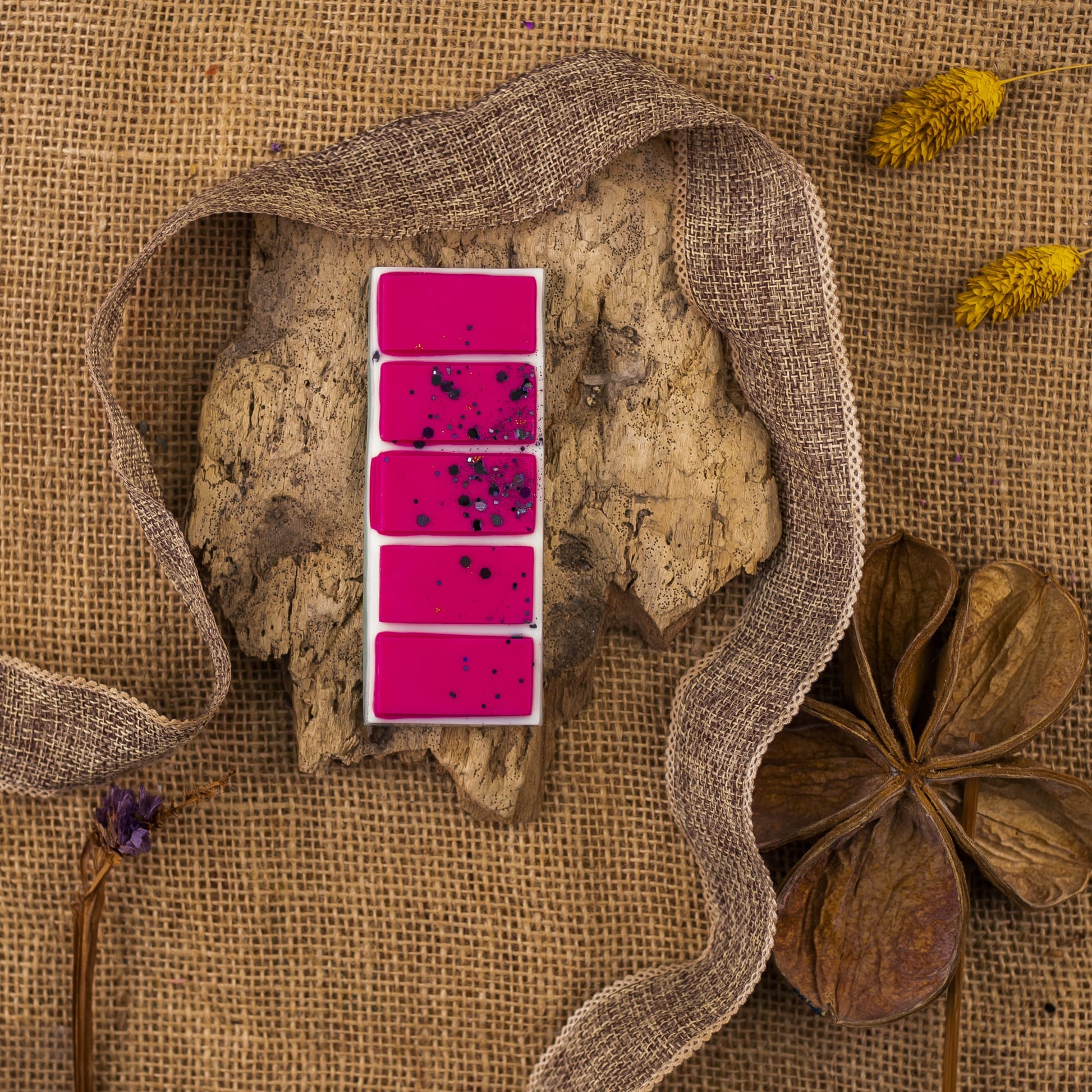Una paleta rectangular de Fleur-de-Bois Wax Melts Láminas Aromáticas DULZURA DE SANDÍA, de color rosa brillante con motas oscuras, descansa sobre una corteza entre arpilleras, flores secas y una vaina de semillas marrones, evocando elegantes fragancias.