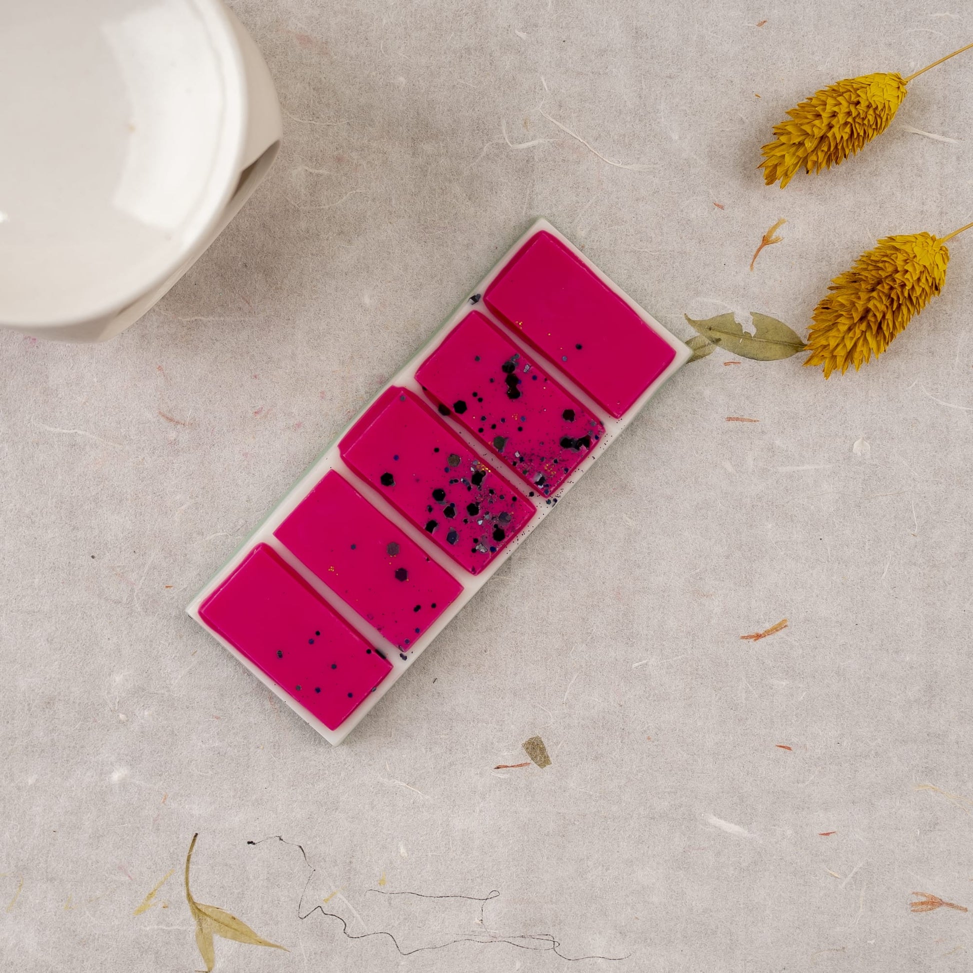Cinco Fusiones de Cera Láminas Aromáticas Fleur-de-Bois DULZURA DE SANDÍA de Fleur-de-Bois de color rosa brillante, algunas con motas negras, en un plato rectangular cerca de flores secas amarillas y una pieza de cerámica blanca, evocan fragancias elegantes.