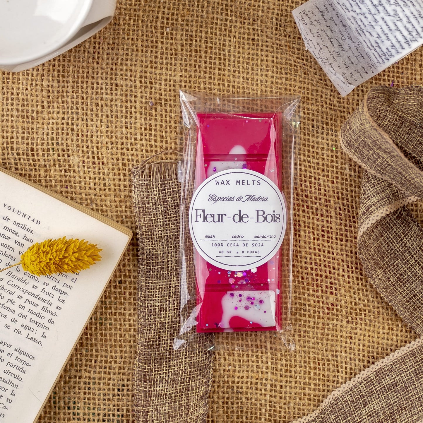 Un paquete de Fleur-de-Bois Wax Melts Láminas Aromáticas ESPECIAS DE MADERA con un diseño rosa y blanco descansa sobre arpillera, rodeado de un lazo, un libro abierto, una flor amarilla seca, notas escritas a mano y fragancias premium cera aromática.