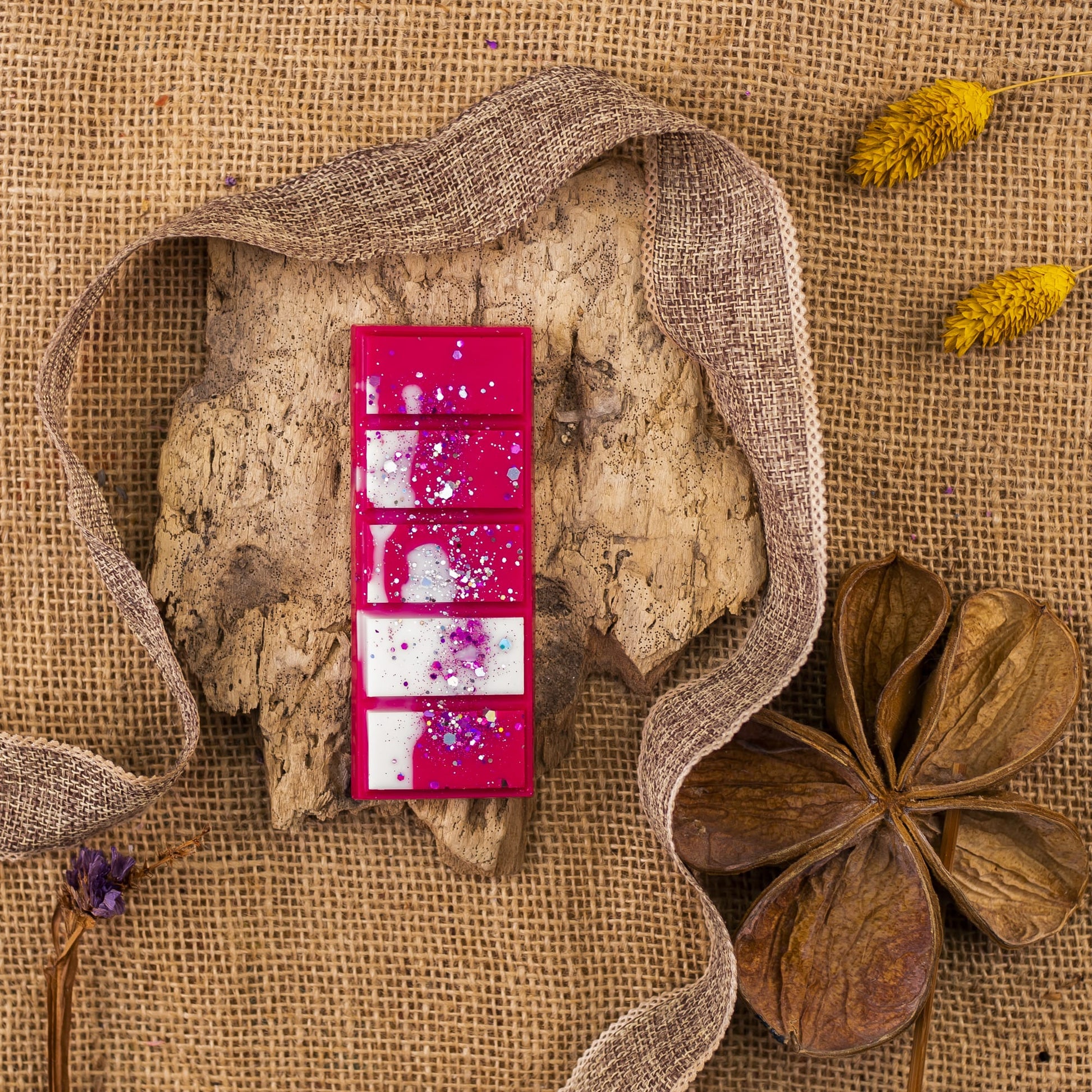 Una paleta de pintura rosa con cinco pocillos, algunos de los cuales contienen Wax Melts morados y blancos Láminas Aromáticas Fleur-de-Bois ESPECIAS DE MADERA de Fleur-de-Bois, descansa sobre corteza de árbol sobre arpillera, rodeada de flores secas y una cinta marrón.