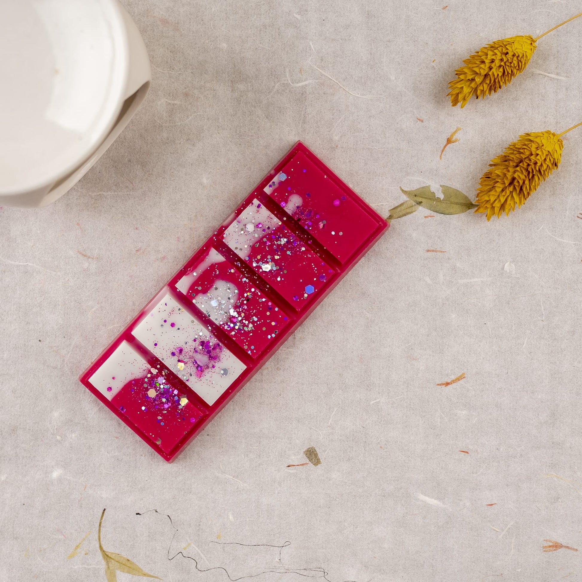 Un rectángulo rosa de Wax Melts Láminas Aromáticas Fleur-de-Bois ESPECIAS DE MADERA de Fleur-de-Bois, dividido en cinco secciones con salpicaduras blancas y moradas, descansa sobre una superficie texturizada cerca de dos flores secas amarillas y un objeto de cerámica blanca.