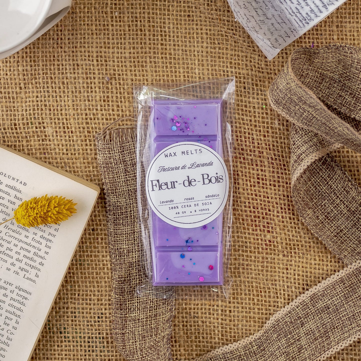Un paquete de Wax Melts Láminas Aromáticas Flor de Bois FRESCURA DE LAVANDA sobre arpillera, con un libro abierto, una flor amarilla seca y una carta cerca, listo para llenar tu espacio de fragancias premium de cera aromática.