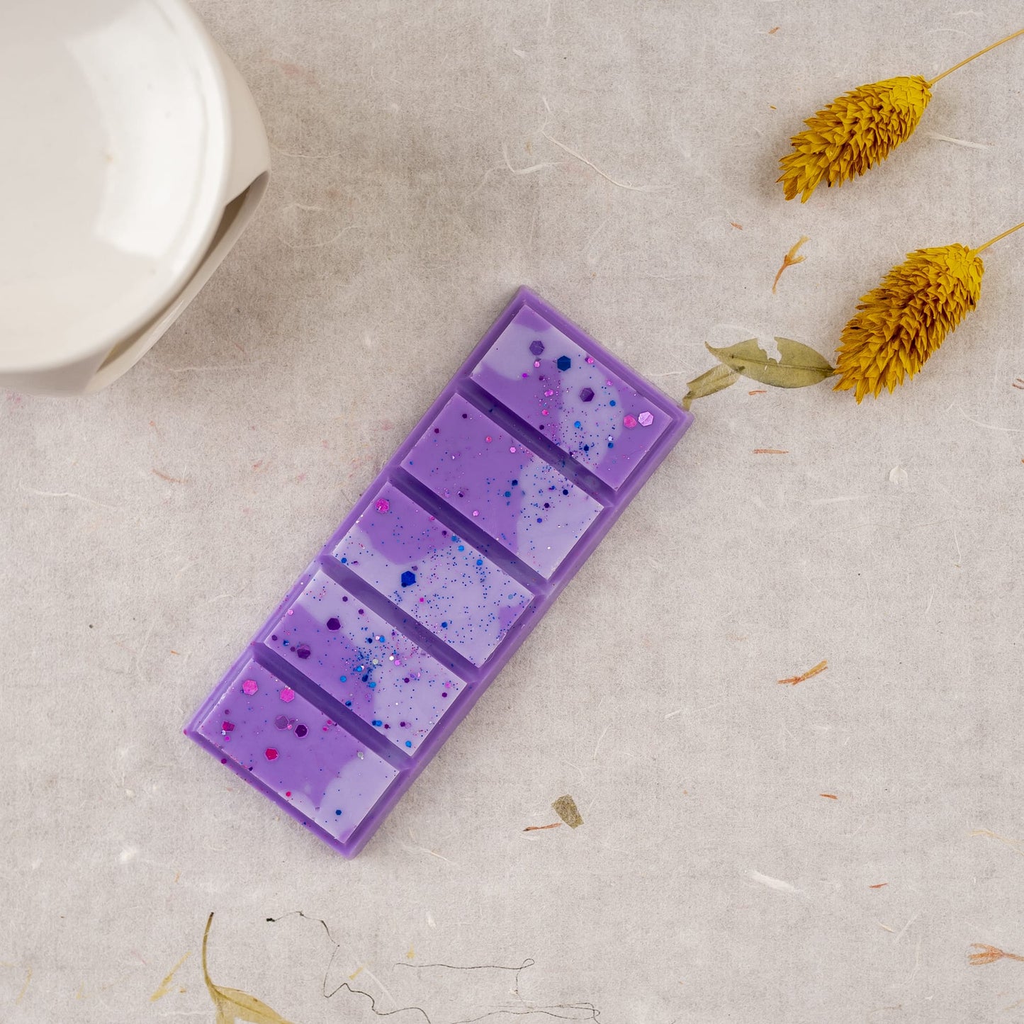 Una barra rectangular morada de Wax Melts Láminas Aromáticas Fleur-de-Bois FRESCURA DE LAVANDA, moteada de azul y rosa, descansa sobre una superficie clara cerca de flores secas amarillas y parte de un objeto de cerámica blanca de Fleur-de-Bois.
