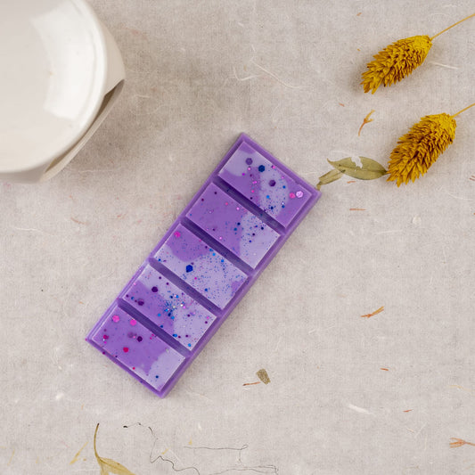 Una barra rectangular morada de Wax Melts Láminas Aromáticas Fleur-de-Bois FRESCURA DE LAVANDA, moteada de azul y rosa, descansa sobre una superficie clara cerca de flores secas amarillas y parte de un objeto de cerámica blanca de Fleur-de-Bois.