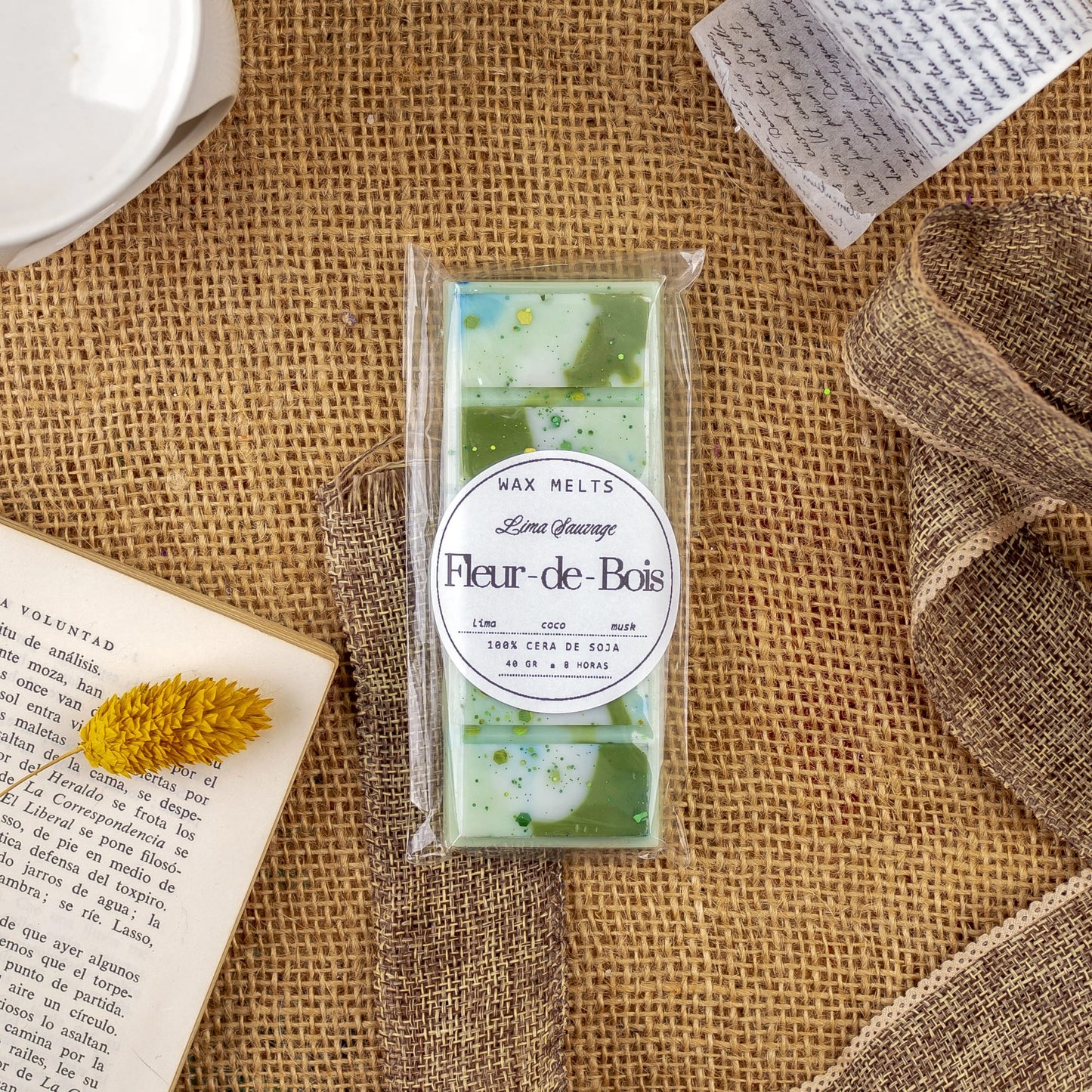 Un paquete de Wax Melts Láminas Aromáticas Fleur-de-Bois LIMA SAUVAGE de Fleur-de-Bois descansa sobre arpillera, rodeado de cinta, un libro abierto, una flor seca y platos, evocando elegantes fragancias.