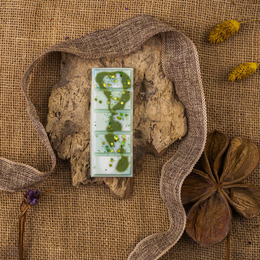 Un plato blanco rectangular con ceras fundidas verdes -Fleur-de-Bois Láminas Aromáticas LIMA SAUVAGE- está colocado sobre corteza de árbol, rodeado de una cinta marrón tejida, flores secas, tela de arpillera y elegantes fragancias que recuerdan a la cera aromática de primera calidad.