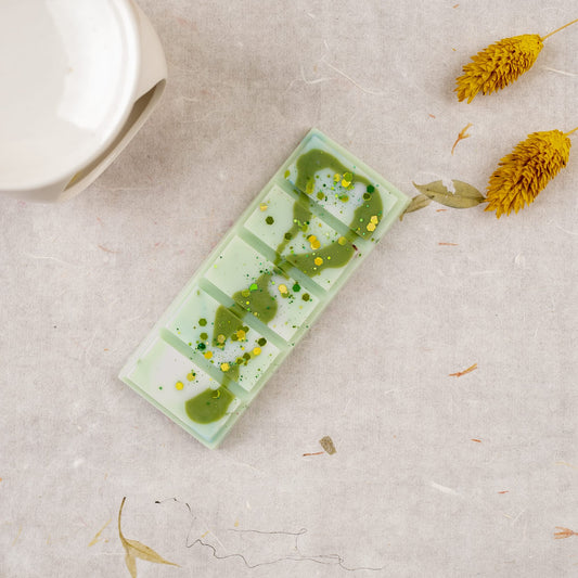 Una paleta rectangular de cerámica verde menta con pintura verde se asienta sobre un beige texturizado, junto a flores amarillas secas y una Flor de Bois Wax Melts Láminas Aromáticas LIMA SAUVAGE, que añaden una elegante fragancia a la escena.