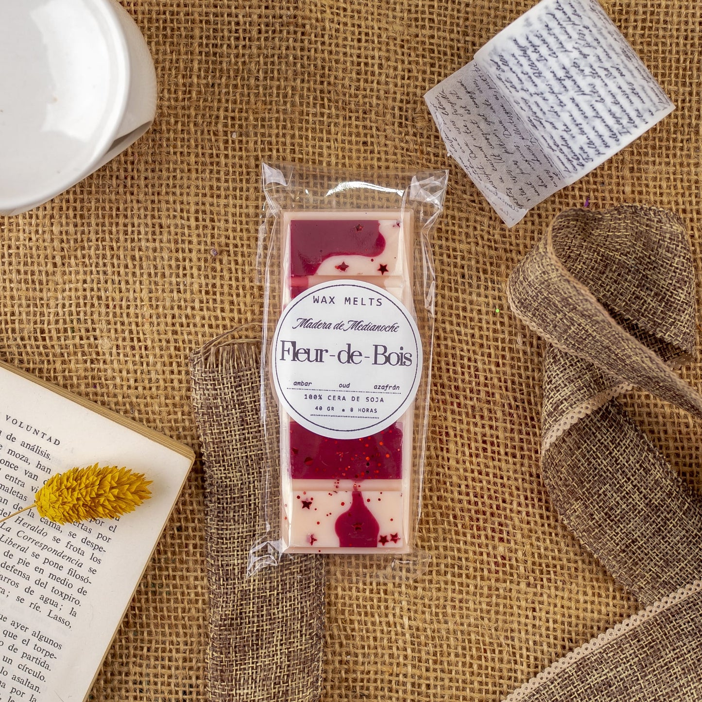 La Cera Fundida Láminas Aromáticas Flor de Bois MADERA DE MEDIANOCHE de Fleur-de-Bois descansa sobre arpillera, rodeada de un libro, una carta manuscrita, una flor seca amarilla y una pieza de cerámica blanca: un encantador despliegue de fragancias de primera calidad.