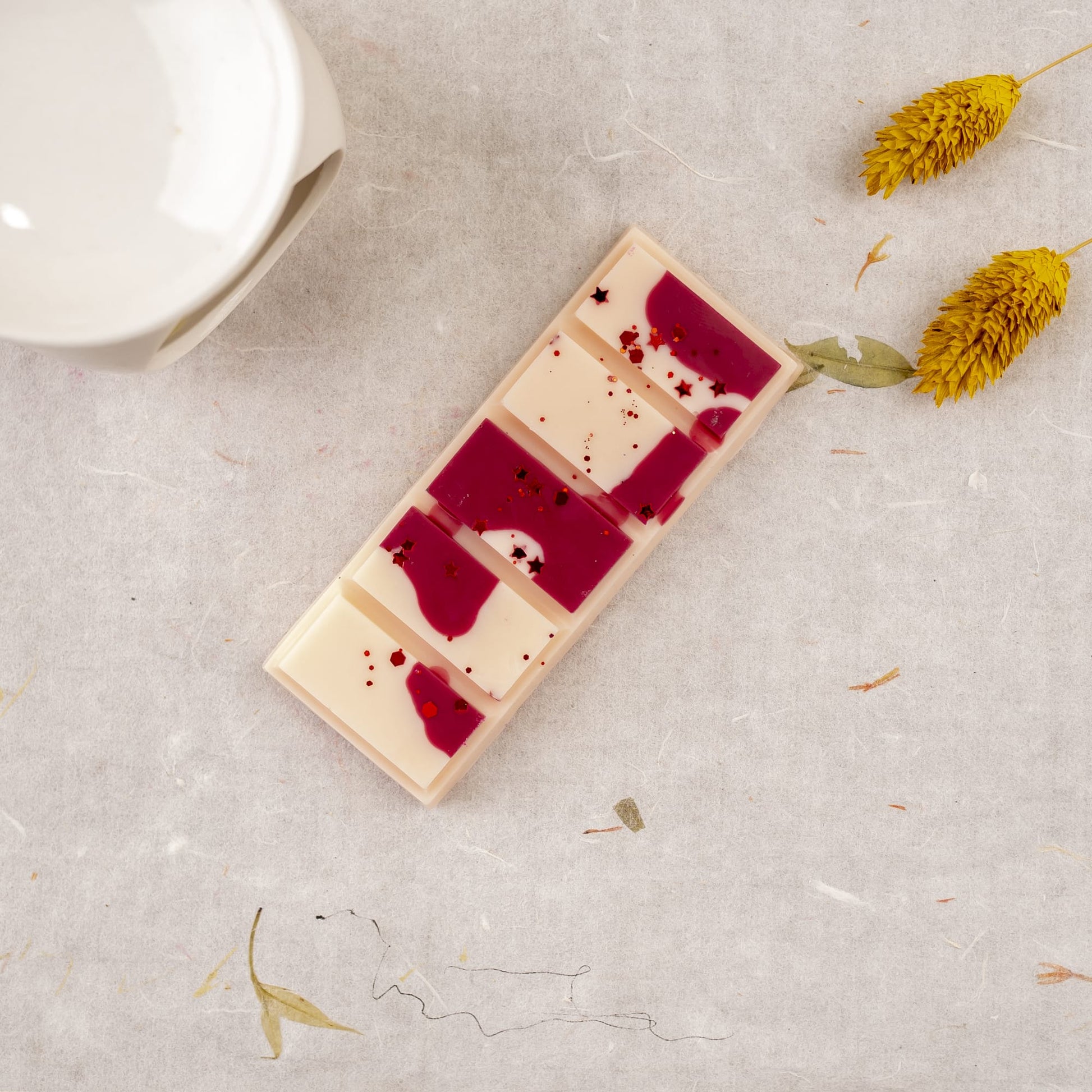 Una tableta rectangular de chocolate blanco y rojo con virutas descansa sobre una superficie texturizada cerca de un recipiente de cerámica blanca, dos flores secas amarillas y Flor de Bois Wax Melts Láminas Aromáticas MADERA DE MEDIANOCHE.