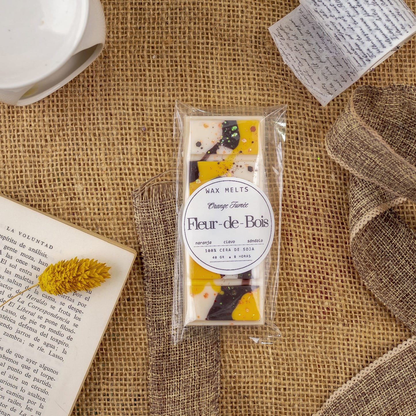 Un paquete de Melts de Cera Láminas Aromáticas Fleur-de-Bois ORANGE FUMÉE de Fleur-de-Bois descansa sobre arpillera, rodeado de una cinta marrón, una flor seca amarilla, un libro abierto, una taza blanca y una nota manuscrita.