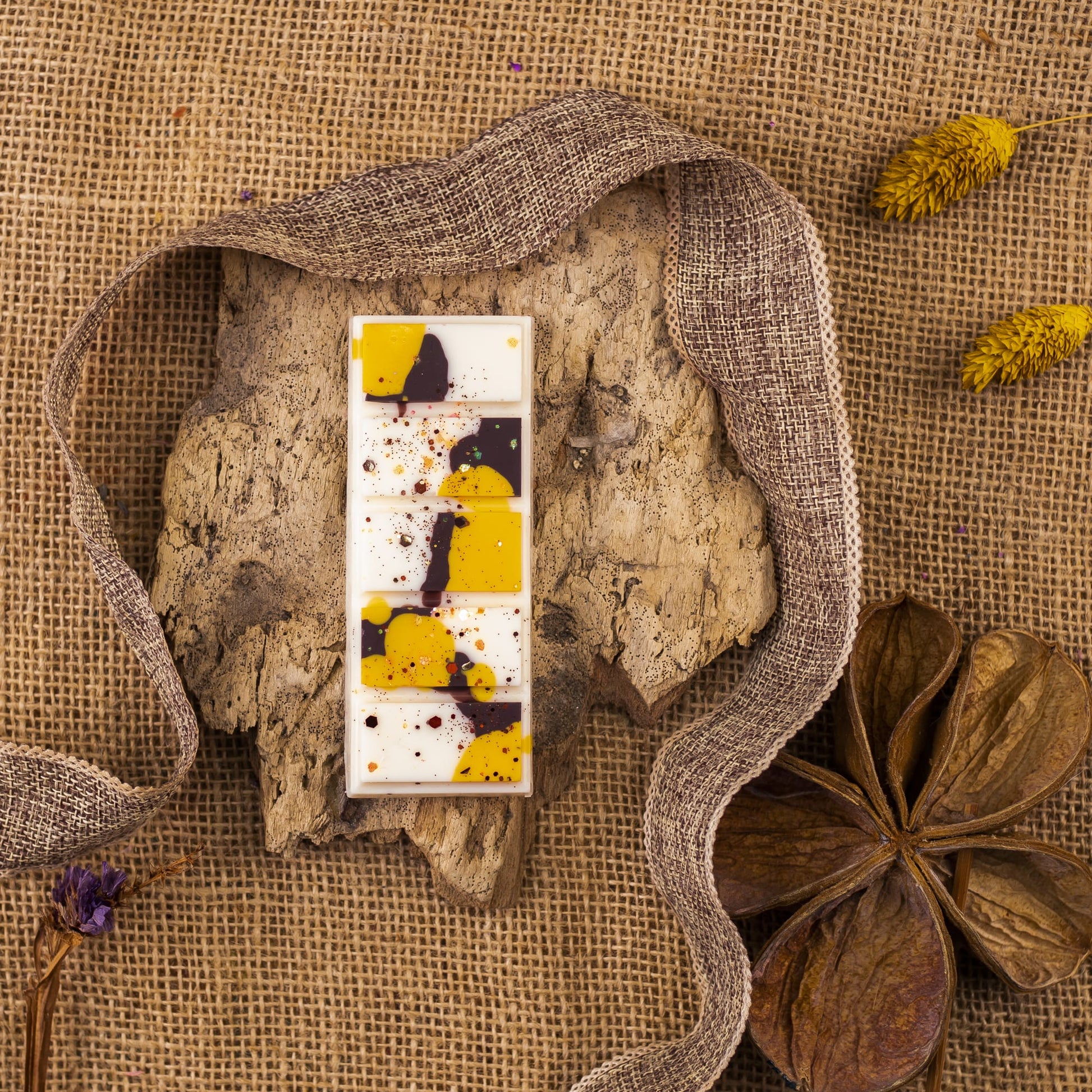 Wax Melts Láminas Aromáticas Fleur-de-Bois ORANGE FUMÉE de Fleur-de-Bois, con salpicaduras amarillas y moradas, descansan sobre una corteza texturizada entre arpillera, una cinta marrón, flores secas y una vaina de semillas, evocando fragancias artesanales de lujo.