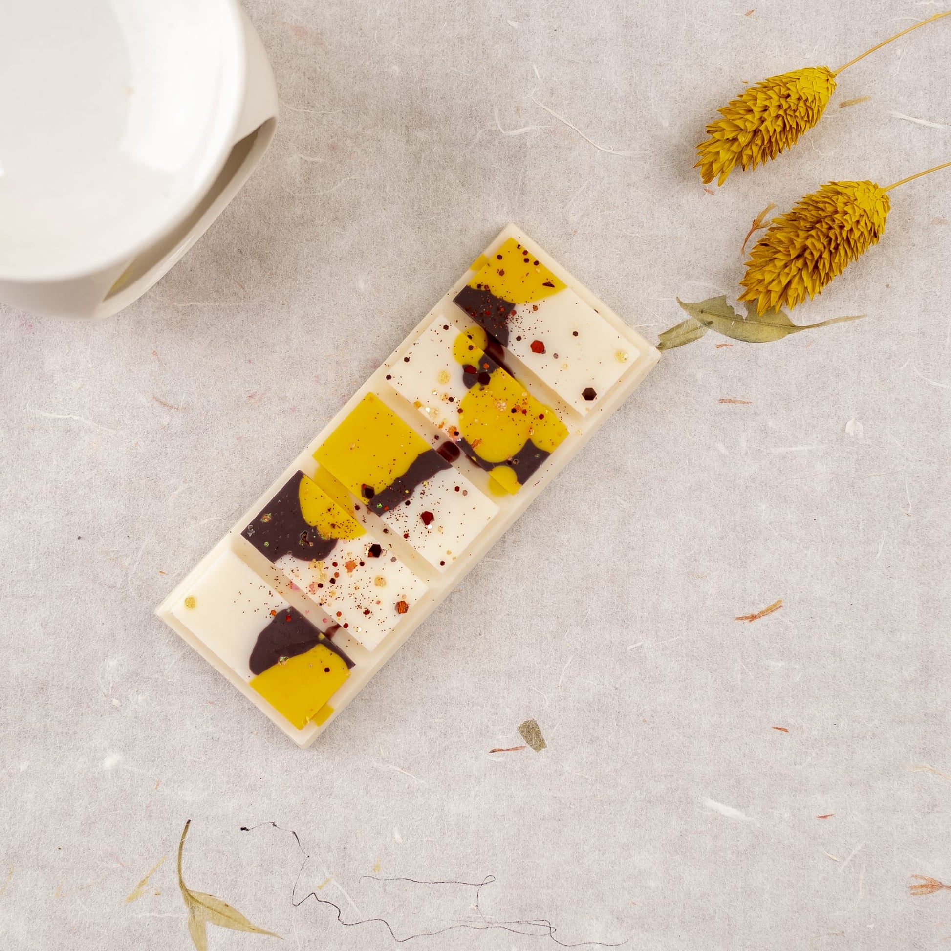 La Cera Fundida Fleur-de-Bois Láminas Aromáticas NARANJA FUMÉE, con remolinos abstractos amarillos y marrones, descansa sobre una superficie de textura ligera junto a una pieza de cerámica blanca y flores amarillas secas, que evocan colecciones de fragancias de primera calidad.