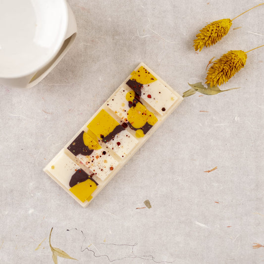 La Cera Fundida Fleur-de-Bois Láminas Aromáticas NARANJA FUMÉE, con remolinos abstractos amarillos y marrones, descansa sobre una superficie de textura ligera junto a una pieza de cerámica blanca y flores amarillas secas, que evocan colecciones de fragancias de primera calidad.