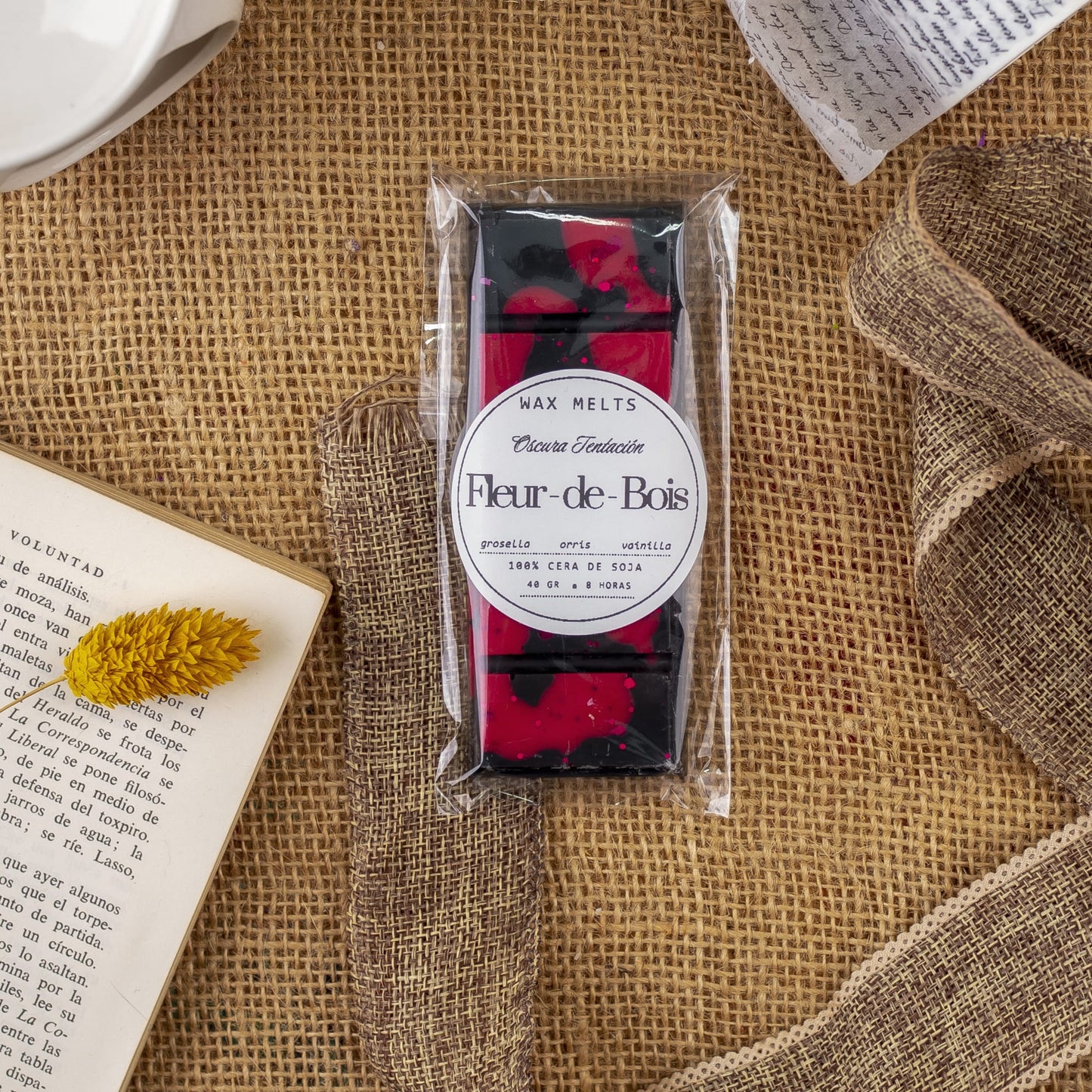 Un paquete de Fleur-de-Bois Wax Melts Láminas Aromáticas OSCURA TENTACIÓN descansa sobre arpillera, rodeado de un libro abierto, una flor amarilla seca, una cinta marrón y papel escrito a mano. La cera negra y roja descansa en un recipiente de plástico transparente.