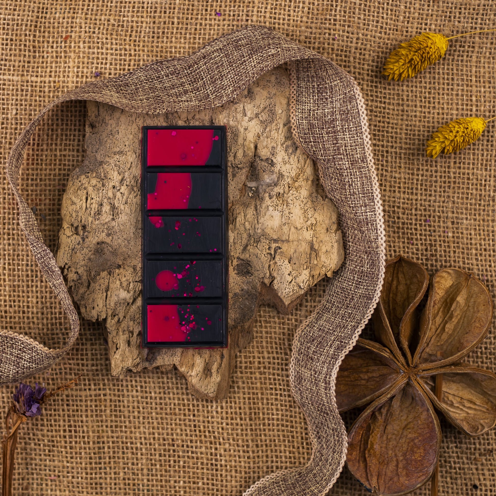 Cuatro Melts de Cera OSCURA TENTACIÓN Fleur-de-Bois rectangulares con salpicaduras rojas descansan sobre madera, rodeados de arpillera, cinta, flores secas amarillas, una vaina en forma de estrella y láminas aromáticas para darle un toque acogedor.