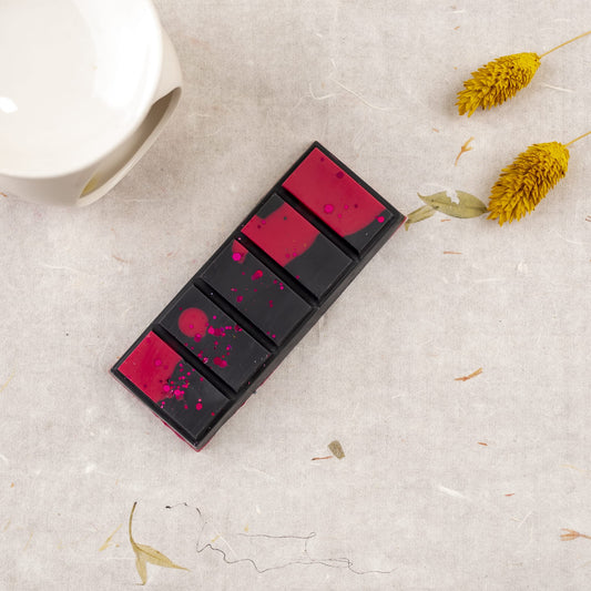 Una barra rectangular de Fleur-de-Bois OSCURA TENTACIÓN Wax Melts Láminas Aromáticas, con brillantes salpicaduras rosas, descansa sobre tela clara junto a una taza de cerámica blanca y dos flores secas amarillas.