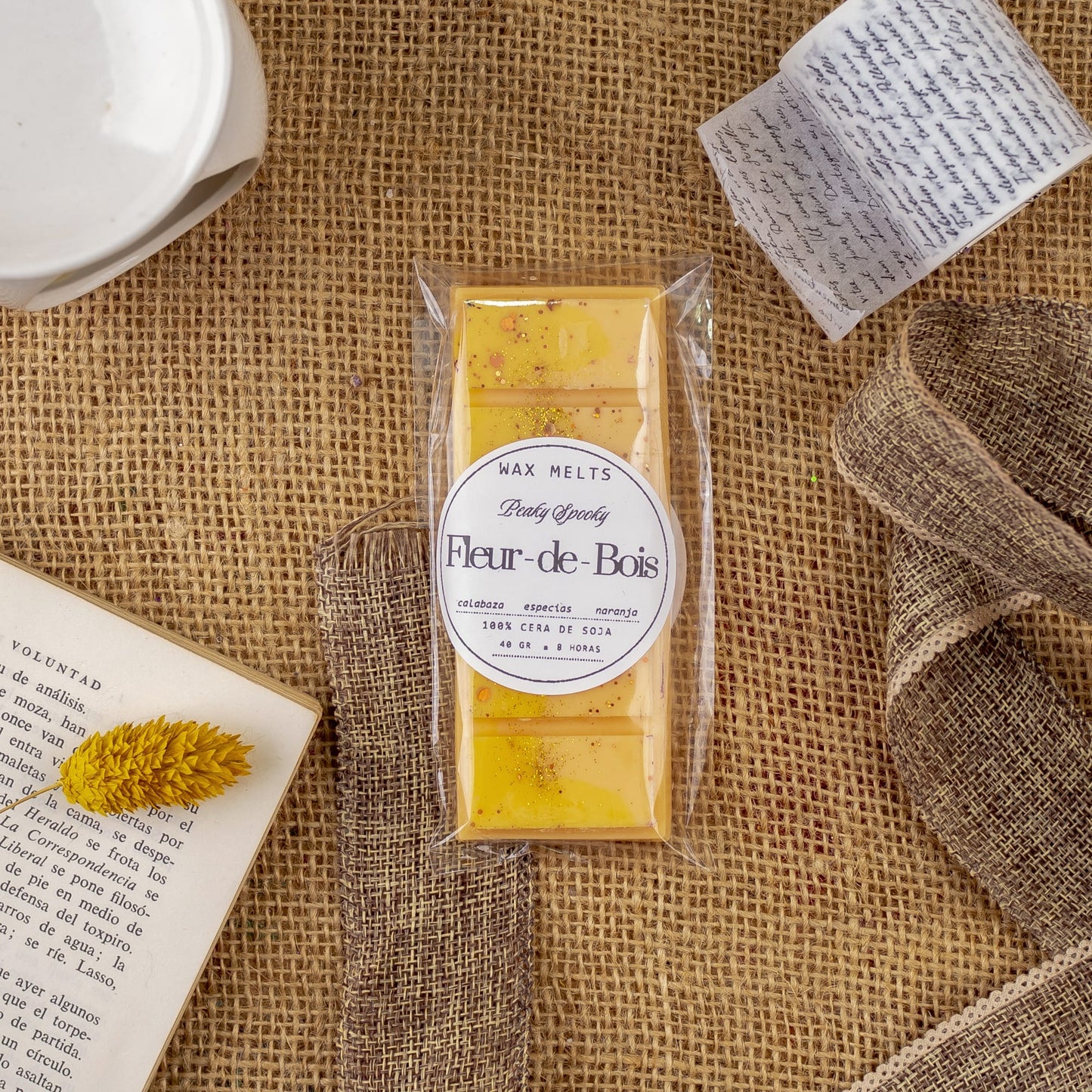 Un paquete de Melts de Cera Láminas Aromáticas Fleur-de-Bois PEAKY SPOOKY de Fleur-de-Bois descansa sobre arpillera junto a un libro abierto, una nota manuscrita, una flor amarilla seca y un lazo, ideal para disfrutar de fragancias caseras de primera calidad.