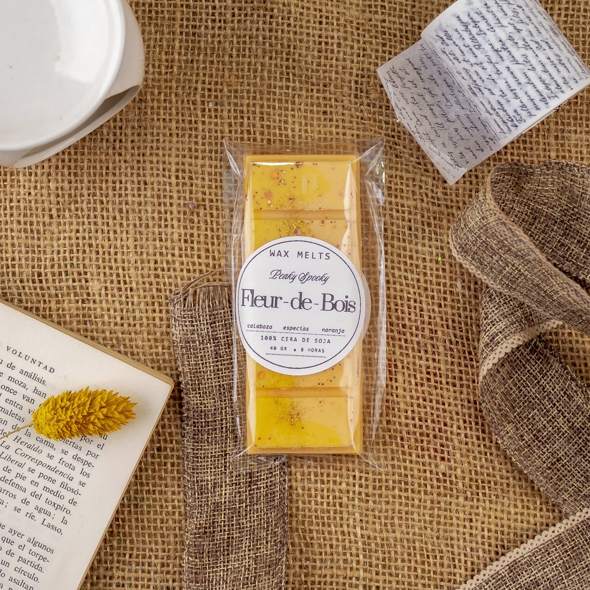 Un paquete de Melts de Cera Láminas Aromáticas Fleur-de-Bois PEAKY SPOOKY de Fleur-de-Bois descansa sobre arpillera junto a un libro abierto, una nota manuscrita, una flor amarilla seca y un lazo, ideal para disfrutar de fragancias caseras de primera calidad.