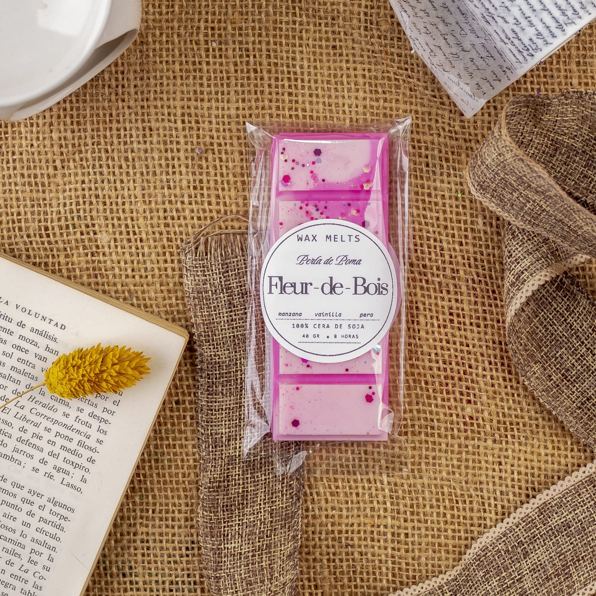 Un paquete de Fleur-de-Bois Wax Melts Láminas Aromáticas PERLA DE POMA descansa sobre arpillera, rodeado de una flor seca amarilla, libros abiertos y papel escrito a mano, evocando elegantes fragancias.