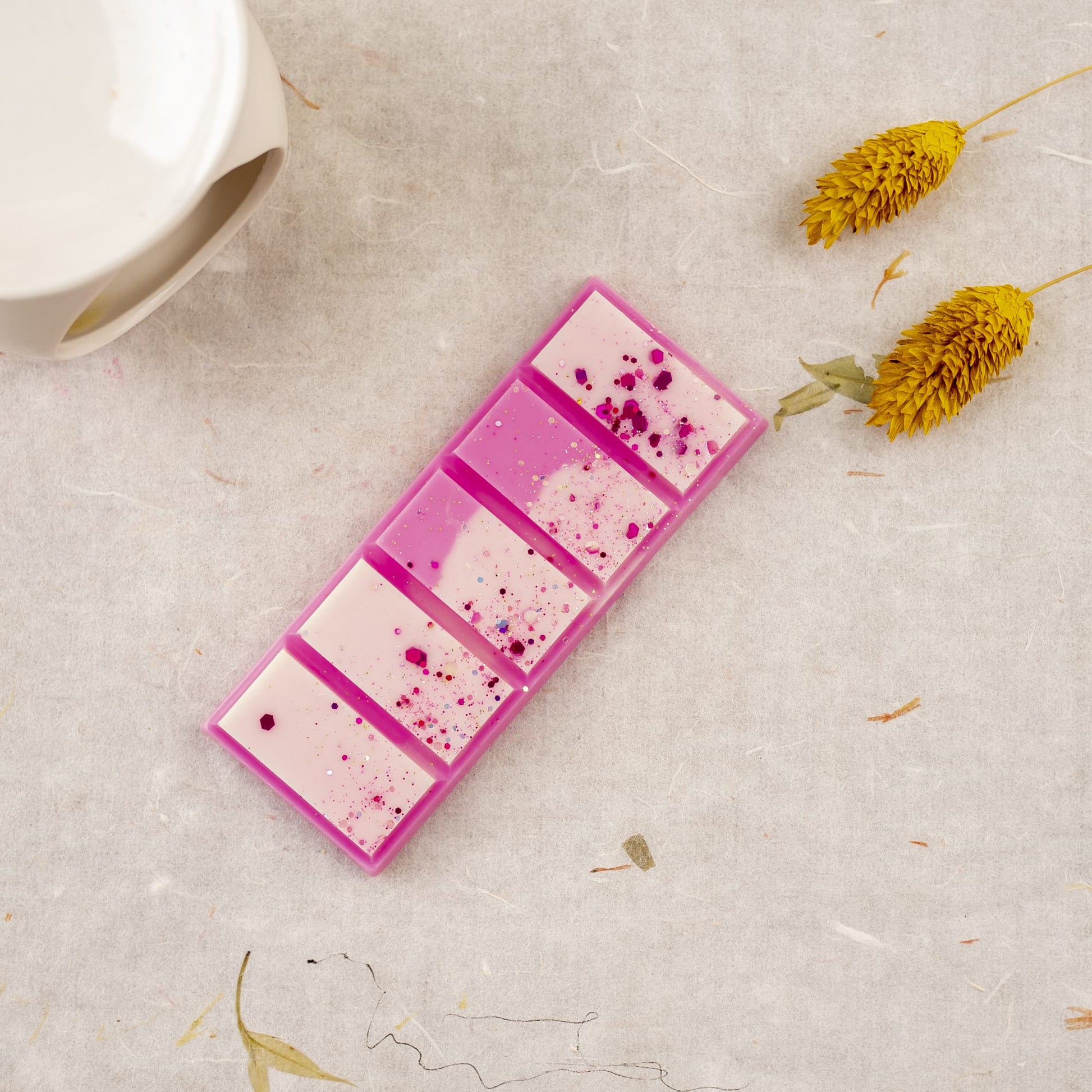 Un rectángulo rosa y blanco Fleur-de-Bois Wax Melts Láminas Aromáticas PERLA DE POMA descansa sobre una superficie texturizada cerca de flores secas amarillas y una cera aromática de cerámica blanca, evocando fragancias elegantes.