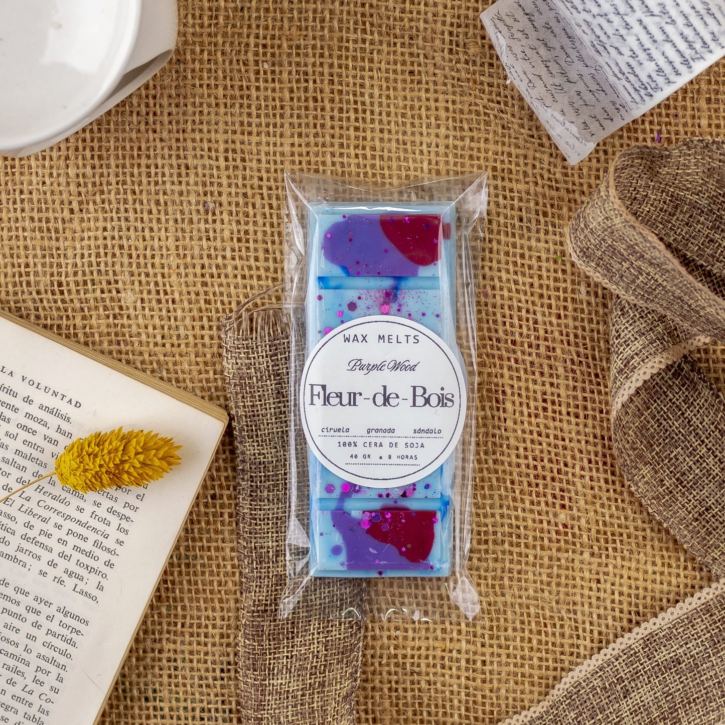Un paquete de Wax Melts de Cera Láminas Aromáticas Fleur-de-Bois PURPLE WOOD de Fleur-de-Bois descansa sobre arpillera junto a un libro abierto, una flor amarilla seca, un lazo y una nota, perfecto para saborear fragancias elegantes.