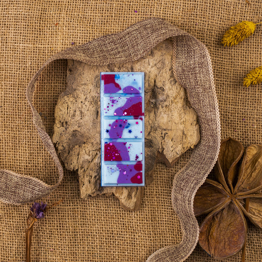 Una hoja de Wax Melts Láminas Aromáticas Fleur-de-Bois PURPLE WOOD de Fleur-de-Bois descansa sobre corteza, con arpillera, una cinta marrón y flores secas, evocando elegantes aromas y un refinado estilo de cera aromática.