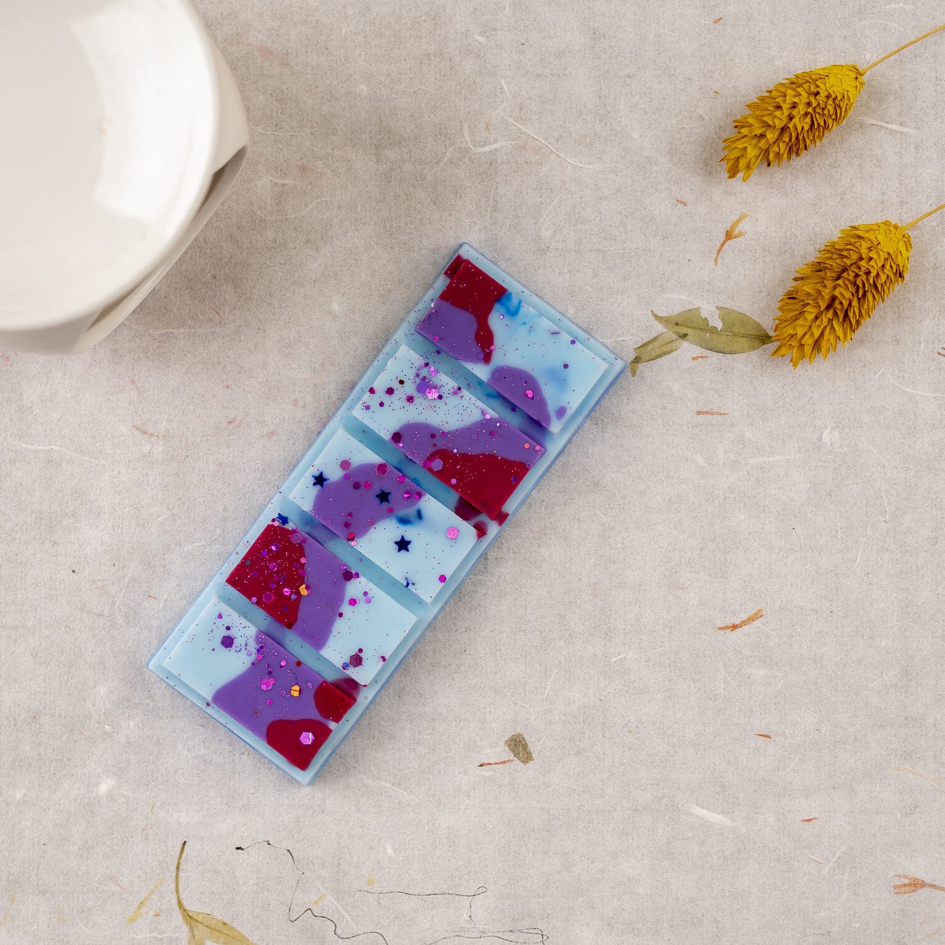 Una barra rectangular de Wax Melts Láminas Aromáticas Fleur-de-Bois PURPLE WOOD con remolinos y purpurina azules, morados y rojos descansa sobre una superficie ligera junto a flores secas amarillas, ofreciendo un aroma personalizado de la marca Fleur-de-Bois para cualquier espacio.