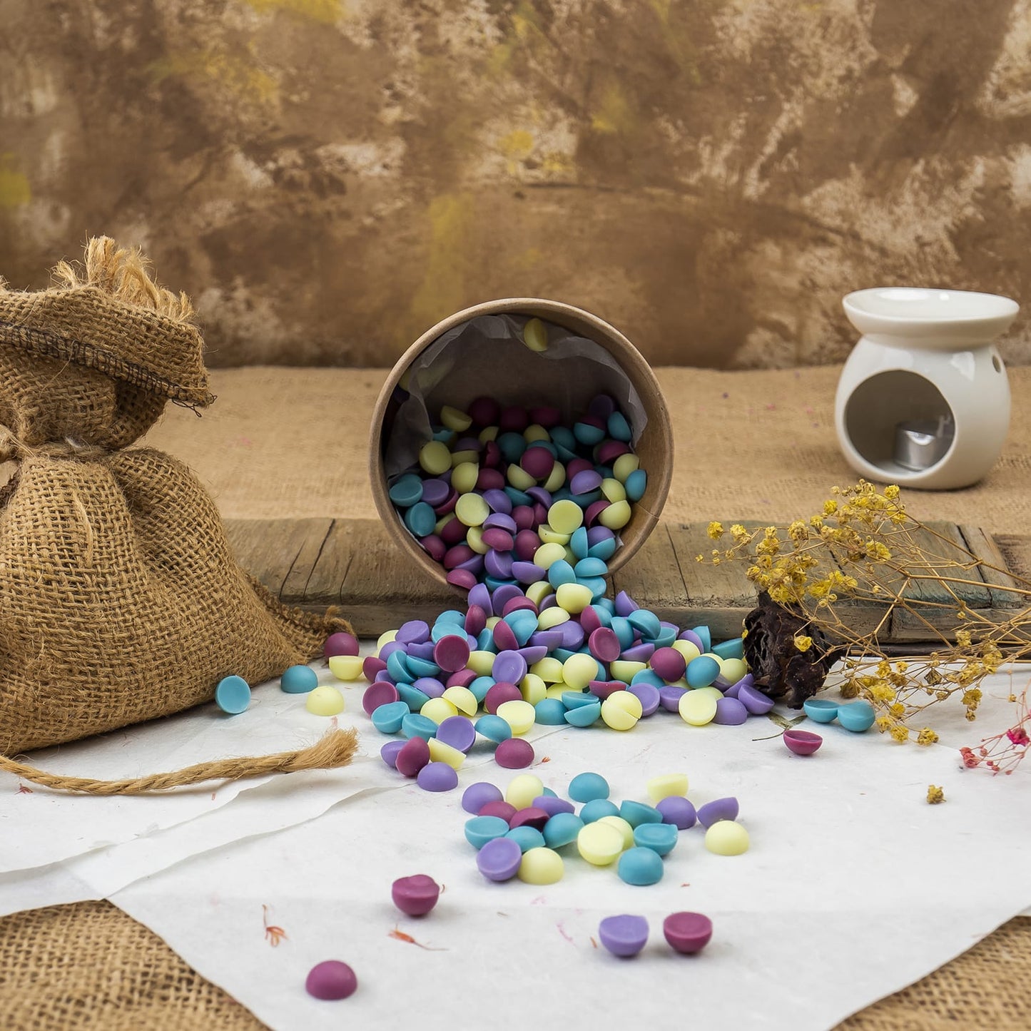 Una taza volcada derrama caramelos redondos de colores sobre madera rústica con arpillera, flores secas y el aroma de Wax Melts Esencias en Miniatura CHISPITAS DE COLORES de Fleur-de-Bois en un quemador de aroma blanco al fondo.