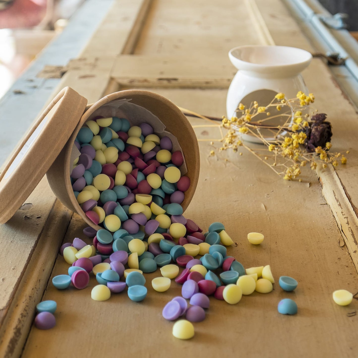 Un recipiente de Esencias en Miniatura CHISPITAS DE COLORES Fleur-de-Bois derrite cera pastel de soja sobre una superficie de madera, con un calentador de cera blanco y flores amarillas secas que realzan el aroma artesanal.