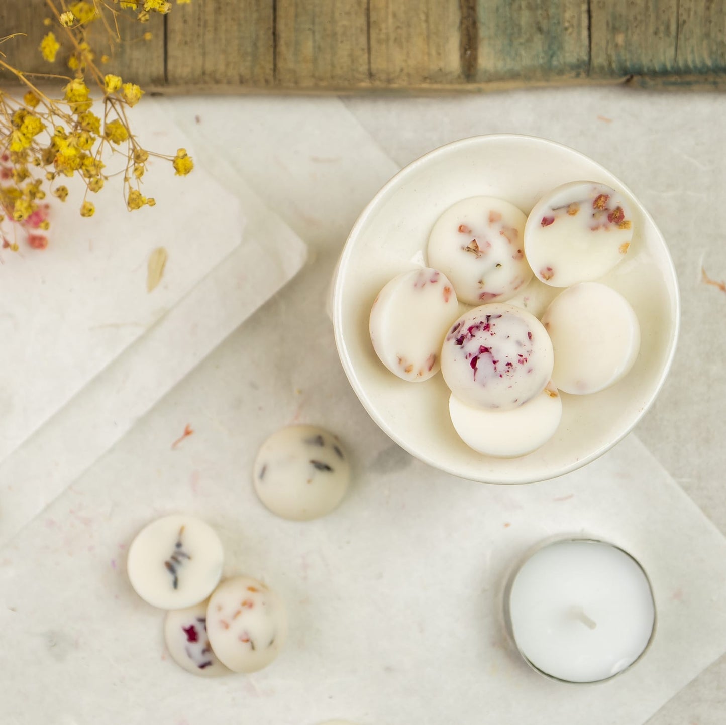 Un cuenco blanco contiene Wax Melts Esencias en Miniatura COPOS DE AROMA de Fleur-de-Bois, rellenos de flores secas. Las Wax Melts esparcidas, una candelita y ramitas secas cercanas lo hacen ideal para disfrutar de las fragancias otoñales esta temporada.