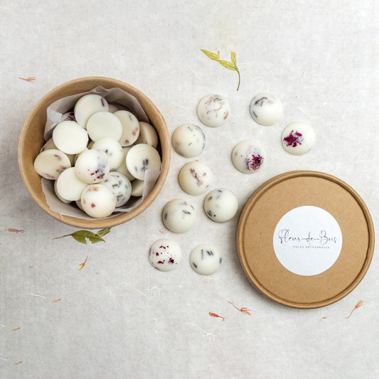 Cera blanca redonda Esencias en Miniatura COPOS DE AROMA de Fleur-de-Bois, decorada con pétalos de flores secas, dentro y alrededor de un recipiente de carton marrón con la etiqueta "Fleur-de-Bois Velas Artesanales", ideal para añadir fragancias otoñales.