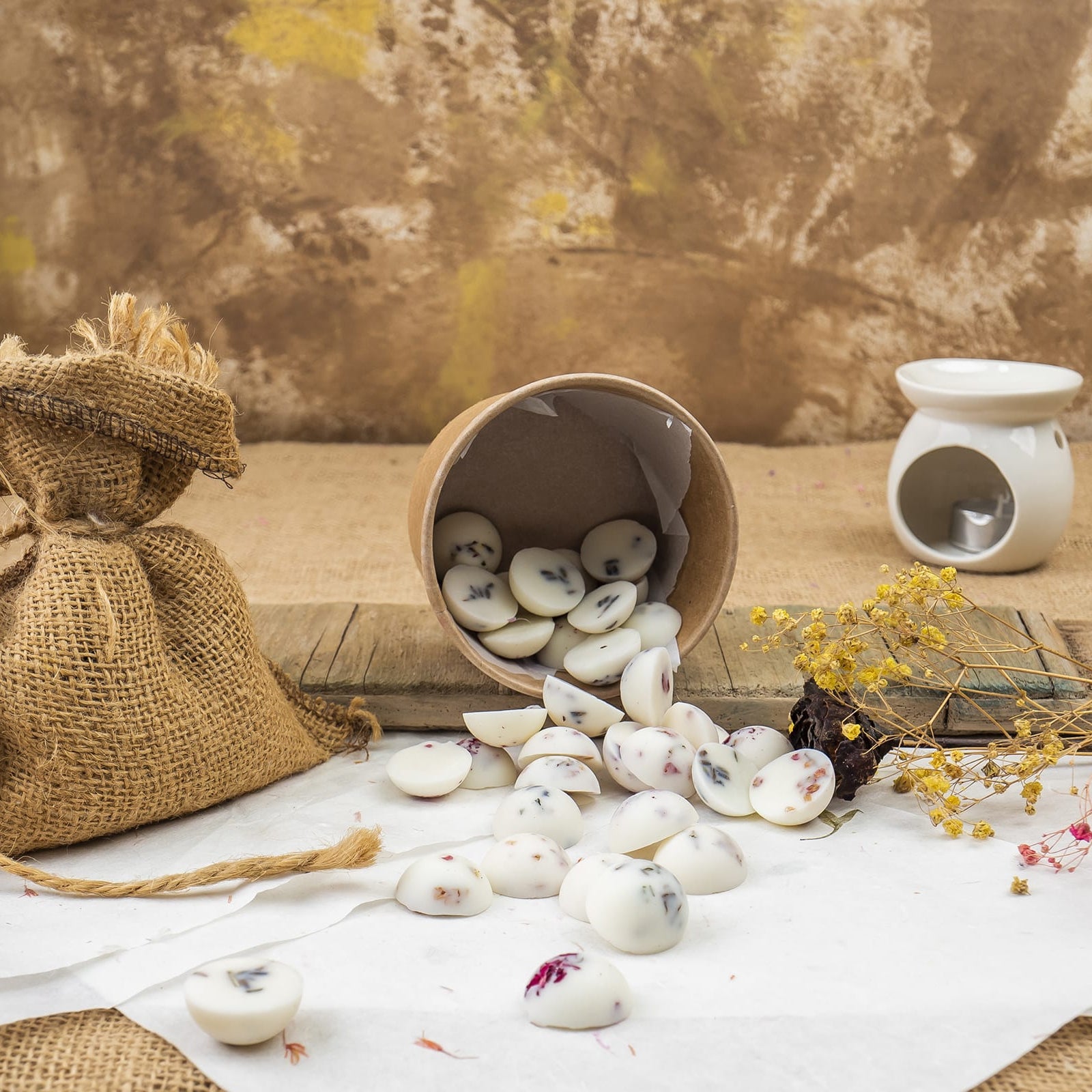 Un vaso de carton marrón derrama Fleur-de-Bois Wax Melts Esencias en Miniatura COPOS DE AROMA con pétalos de flores secas. Cerca, una bolsa de arpillera, flores secas y un calentador de cera fundida blanca descansan sobre una superficie rústica contra un fondo marrón texturizado.