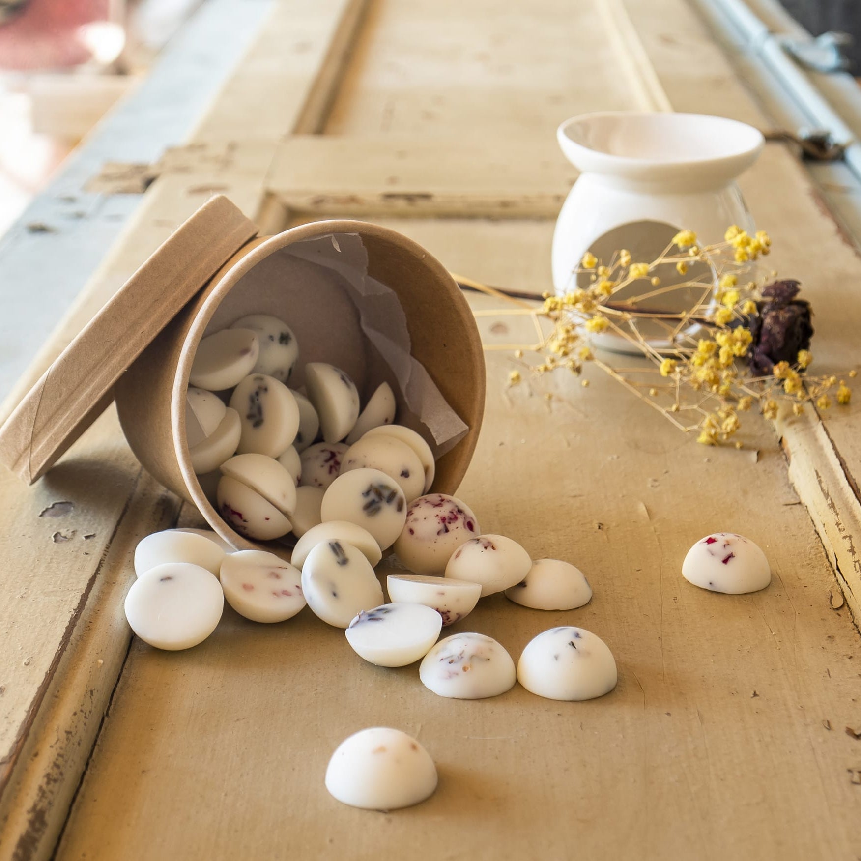 Un recipiente de carton marrón derrama Wax Melts Esencias en Miniatura COPOS DE AROMA de Fleur-de-Bois y pétalos de flores sobre madera rústica. Un quemador de cera y flores amarillas secas en el fondo añaden un toque acogedor y aromático.