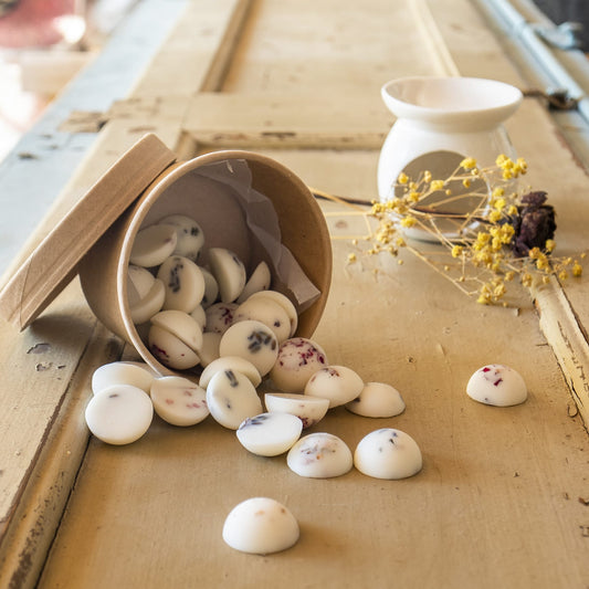 Un recipiente de carton marrón derrama Wax Melts Esencias en Miniatura COPOS DE AROMA de Fleur-de-Bois y pétalos de flores sobre madera rústica. Un quemador de cera y flores amarillas secas en el fondo añaden un toque acogedor y aromático.